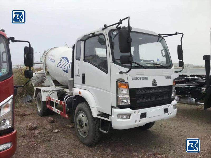 Vue latérale d'un camion malaxeur à béton HOWO 4x2 blanc garé sur un terrain en gravier, montrant la cabine du conducteur et le tambour de malaxage cylindrique avec la marque de l'entreprise.